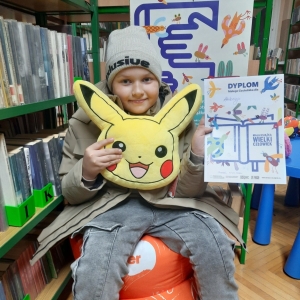 W bibliotece. Uśmiechnięty chłopiec siedzi na pomarańczowej pufie. W ręku trzyma dyplom i pluszową poduszkę w kolorze żółtym. W tle regały z książkami.