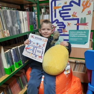 W bibliotece. Uśmiechnięty chłopiec siedzi na pomarańczowej pufie. W ręku trzyma dyplom i pluszową poduszkę w kolorze żółtym. W tle regały z książkami.