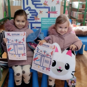 W bibliotce. Dwie dziewczynki siedzą na niebieskich krzesłach. Trzymają dyplomy i pluszowe maskotki.  W tle regały z książkami.