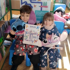 W bibliotce. Dwoje dzieci siedzi na niebieskich krzesłach. Trzymają dyplomy i pluszowe maskotki. W tle regały z książkami.