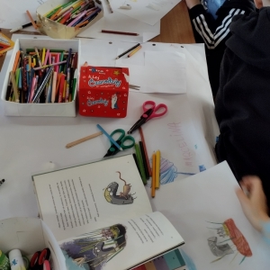 W bibliotece. Na stolikach rozłożone białe kartki, kolorowe kredki i inne przybory potrzebne do wykonania plastycznych prac.