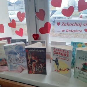 W bibliotece. Na parapecie stoją książki. Na oknie przyklejone czerwone serduszka i napis "Zakochaj się w książce".
