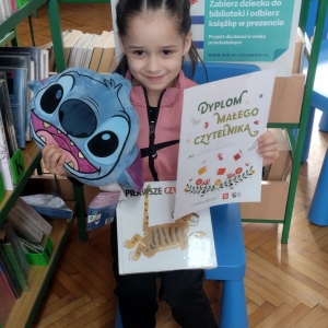 W bibliotece. Uśmiechnięta dziewczynka siedzi na niebieskim krześle. W ręku trzyma dyplom i pluszową poduszkę w kolorze niebieskim. W tle regały z książkami.