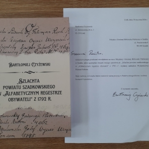 Na stoliku leży książka. Obok niej biała kartka z informacją o przekazaniu książki do biblioteki.