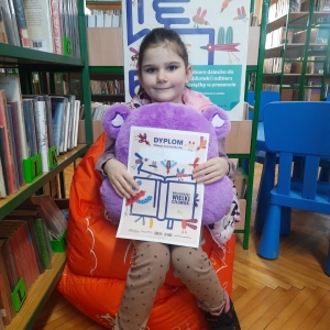 W bibliotece. Uśmiechnięta dziewczynka siedzi na pomarańczowej pufie. W ręku trzyma dyplom i pluszową poduszkę w kolorze fioletowym. W tle regały z książkami