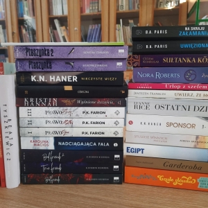 W bibliotece. Na stoliku ustawione książki. W tle regały z książkami.