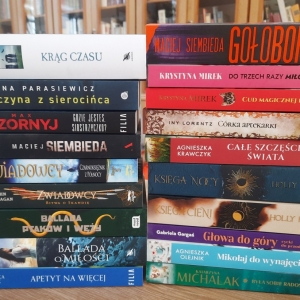 W bibliotece. Na stoliku ustawione książki. W tle regały z książkami.