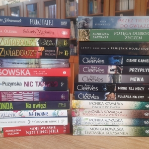 W bibliotece. Na stoliku ustawione książki. W tle regały z książkami.