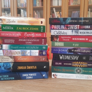 W bibliotece. Na stoliku ustawione książki. W tle regały z książkami.