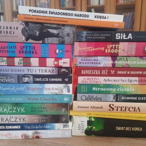 W bibliotece. Na stoliku ustawione książki. W tle regały z książkami.