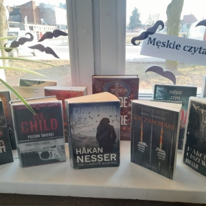 W bibliotece. Na parapecie stoją książki. Na oknie przyklejone papierowe wąsy i napis "Męskie czytanie".