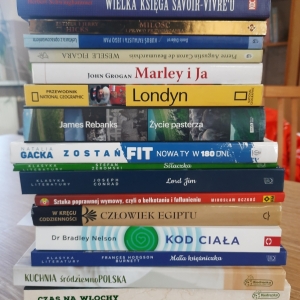 W bibliotece. Na stoliku ułożone książki. W tle regały z książkami.