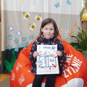 W bibliotece. Uśmiechnięta dziewczynka siedzi na pomarańczowej pufie. W ręku trzyma dyplom.