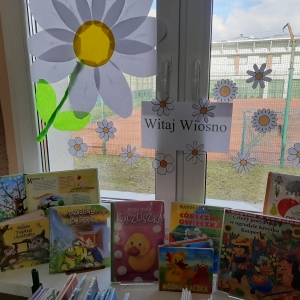 W bibliotece. Na parapecie stoją książki. Na oknie przyklejone papierowe kwiaty i napis "Witaj Wiosno"..