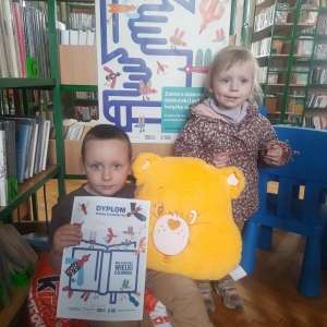 W bibliotece. Dwoje dzieci ustawiło się do zdjęcia. W tle regały z książkami.