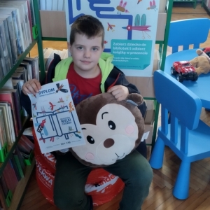 W bibliotece. Uśmiechnięty chłopiec siedzi na pomarańczowej pufie. W ręku trzyma dyplom i pluszową poduszkę w kolorze brązowym. W tle regały z książkami.