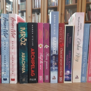 W bibliotece. Na stoliku ustawione książki. W tle regały z książkami.