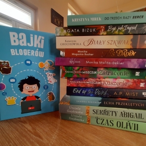 W bibliotece. Na stoliku ustawione książki. W tle regały z książkami.