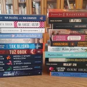 W bibliotece. Na stoliku ustawione książki. W tle regały z książkami.