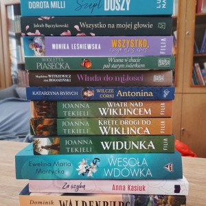 W bibliotece. Na stoliku ustawione książki. W tle regały z książkami.