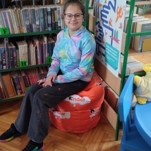 W bibliotece. Uśmiechnięta dziewczynka siedzi na pomarańczowej pufie. W tle regały z książkami.