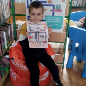 W bibliotece. Uśmiechnięty chłopiec siedzi na pomarańczowej pufie. W ręku trzyma dyplom. W tle regały z książkami.