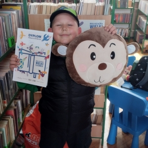 W bibliotece. Uśmiechnięty chłopiec trzyma dyplom i pluszową poduszkę w kolorze brązowym. W tle regały z książkami.