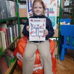 W bibliotece. Uśmiechnięta dziewczynka siedzi na pomarańczowej pufie. W ręku trzyma dyplom. W tle regały z książkami.