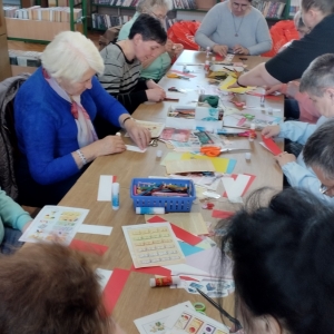 W bibliotece. Podczas warsztatów. Grupa osób siedzi przy stolikach i wykonuje zakładki do książek.