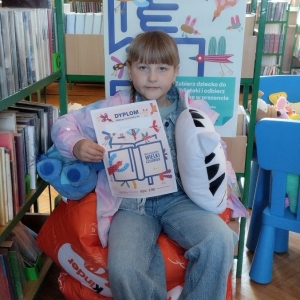 W bibliotece. Dziewczynka siedzi na pomarańczowej pufie. W ręku trzyma dyplom i pluszowe poduszki. W tle regały z książkami.