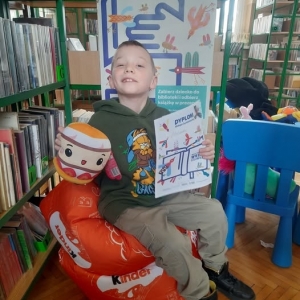 W bibliotece. Uśmiechnięty chłopiec siedzi na pomarańczowej pufie. W ręku trzyma dyplom i maskotkę. W tle regały z książkami.