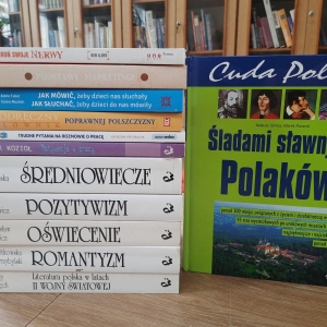 W bibliotece. Na stoliku ustawione książki. W tle regały z książkami.