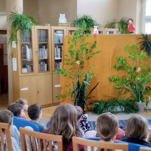 W bibliotece. Dekoracja sceny, miodowa kotara, a na niej zielone rośliny. Nad kotarą kukiełki. Dzieci oglądaja teatrzyk. 