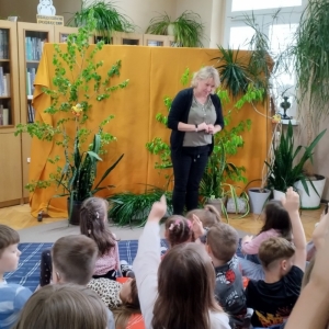 W bibliotece. Dekoracja sceny. Dzieci patrzą w stronę  prowadzącej spotkanie kobiety.