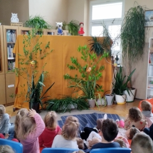 W bibliotece. Scena przygotowana do teatrzyku. Dekoracja to zielone rośliny na miodowym tle.