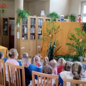 W bibliotece. Dekoracja sceny, miodowa kotara, a na niej zielone rośliny. Nad kotarą kukiełki. Dzieci oglądaja teatrzyk.