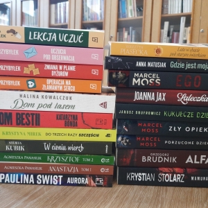 W bibliotece. Na stoliku ustawione książki. W tle regały z książkami.