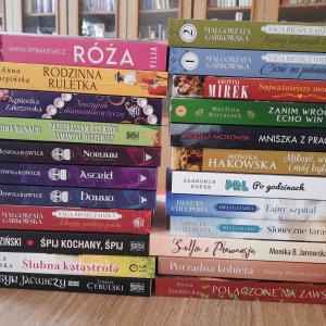 W bibliotece. Na stoliku ustawione książki. W tle regały z książkami.