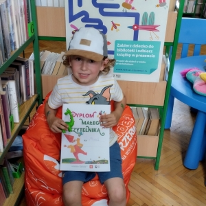 W bibliotece. Chłopiec siedzi na pomarańczowej pufie. W ręku trzyma dyplom. W tle regały z książkami.