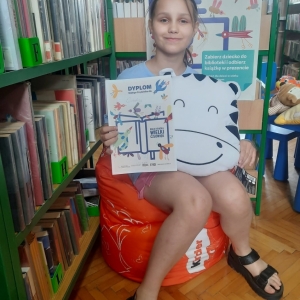W bibliotece. Uśmiechnięta dziewczynka siedzi na pomarańczowej pufie. W ręku trzyma dyplom i pluszową poduszkę. W tle regały z książkami.