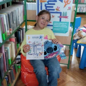 W bibliotece. Uśmiechnięta dziewczynka siedzi na pomarańczowej pufie. W ręku trzyma dyplom i pluszową poduszkę. W tle regały z książkami.
