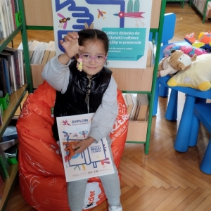 W bibliotece. Uśmiechnięta dziewczynka siedzi na pomarańczowej pufie. W ręku trzyma dyplom i przywieszkę do kluczy - konika Pony. W tle regały z książkami.