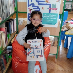 W bibliotece. Uśmiechnięta dziewczynka siedzi na pomarańczowej pufie. W ręku trzyma dyplom. W tle regały z książkami.