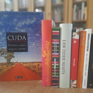W bibliotece. Na stoliku ustawione książki. W tle regały z książkami.