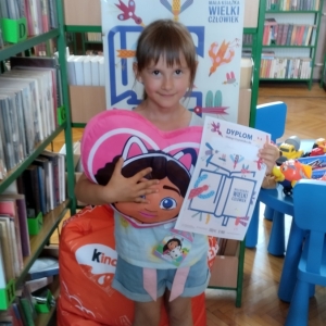 W bibliotece. Uśmiechnięta dziewczynka stoi przy regałach z książkami. W ręku trzyma dyplom i pluszową poduszkę w kolorze różowym.