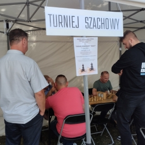 W plenerze. W trakcie trwania turnieju szachowego. Przy stolikach siedzą gracze, obok stoją osoby obserwujące.