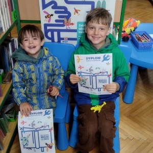 W bibliotce. Dwóch chłopców siedzi na niebieskich krzesłach. Trzymają dyplomy. W tle regały z książkami.