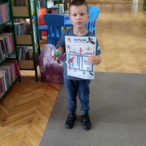 W bibliotece. Chłopiec stoi przy regałach z książkami. W ręku trzyma dyplom i piłkę.