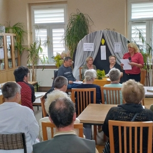 W bibliotece. Grupa osób siedzi na krzesłach. Na fotografii widać trzy osoby siedzące za stołem,  jedna stoi z mikrofonem i kartką. 