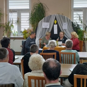W bibliotece. Grupa osób siedzi na krzesłach.  Na fotografii widać cztery osoby siedzące za stołem.